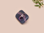 A clear acrylic pin badge with a pink heart-shaped pin back clasp. The design of the pin badge is of a cute chibi penguin dancing in a suit under a spotlight with the quote give 'em the ol' razzle dazzle
