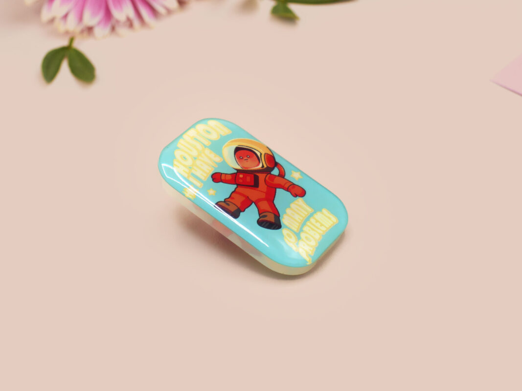 A clear acrylic pin badge with a pink heart-shaped pin back clasp. The design of the pin badge is of a cute chibi retro astronaut floating in sapce with the quote Houston I have so many problems