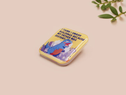 A clear acrylic pin badge with a pink heart-shaped pin back clasp. The design of the pin badge is of a cute chibi blue godzilla like kaiju destroying a city with the quote I Think I Smash Buildings Because My Father Was Distant.