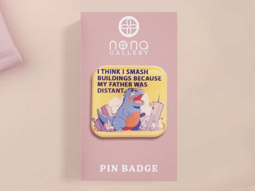 A clear acrylic pin badge with a pink heart-shaped pin back clasp. The design of the pin badge is of a cute chibi blue godzilla like kaiju destroying a city with the quote I Think I Smash Buildings Because My Father Was Distant.