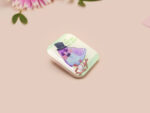 A clear acrylic pin badge with a pink heart-shaped pin back clasp. The design of the pin badge features a cute chibi pigeon wearing a top hat, crying next to a pathetic attempt at nest building, with the quote 'I'm trying my best.'
