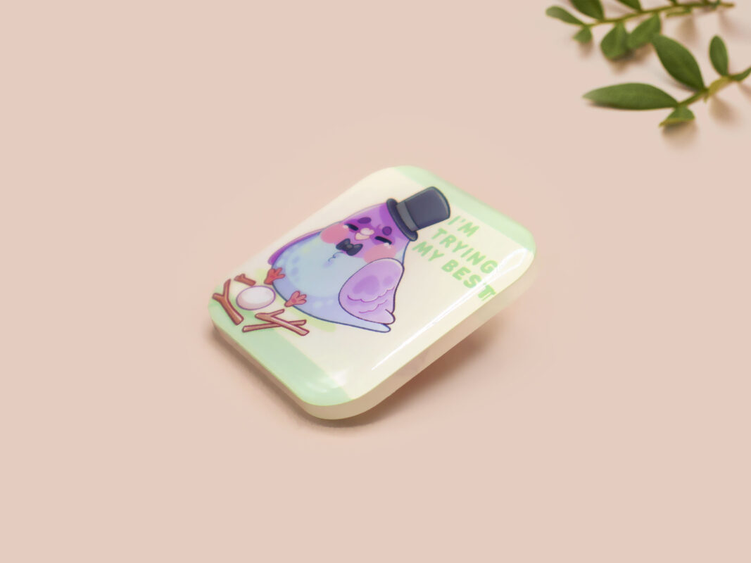 A clear acrylic pin badge with a pink heart-shaped pin back clasp. The design of the pin badge features a cute chibi pigeon wearing a top hat, crying next to a pathetic attempt at nest building, with the quote 'I'm trying my best.'