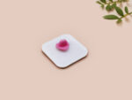 A clear acrylic pin badge with a pink heart-shaped pin back clasp. The design of the pin badge features a cute chibi pigeon wearing a top hat, crying next to a pathetic attempt at nest building, with the quote 'I'm trying my best.'