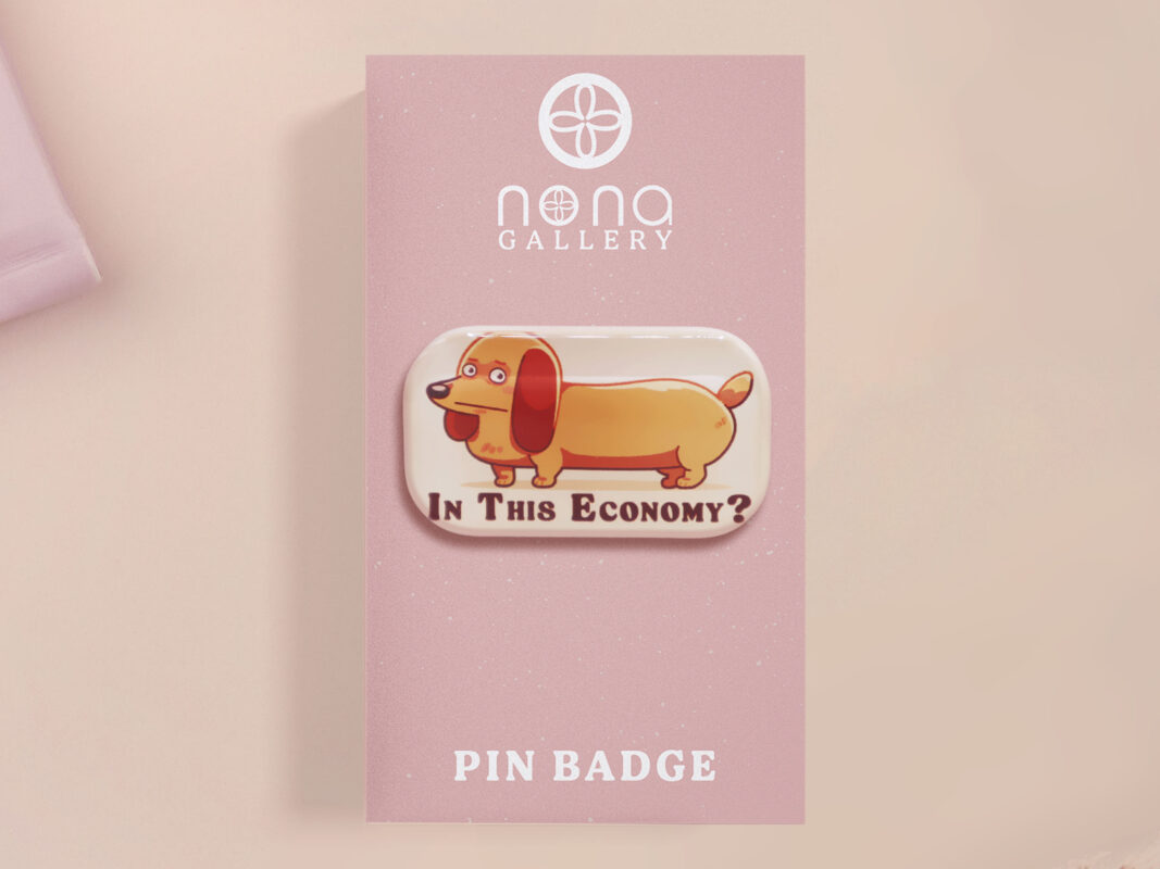 A clear acrylic pin badge with a pink heart-shaped pin back clasp. The design of the pin badge is of a cute chibi dachshund sausage dog with the quote in this economy?
