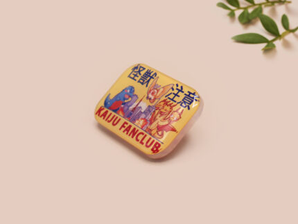 A clear acrylic pin badge with a pink heart-shaped pin back clasp. The design of the pin badge is of a cute chibi blue godzilla like kaiju mothra and King Ghidorah destroying a city with the quote kaiju fanclub and kaiju warning in japanese kanji