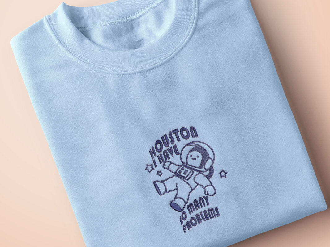 T-shirt with an embroidered blue design of a cute chibi astronaut surrounded by the funny quote Houston I have so many problems