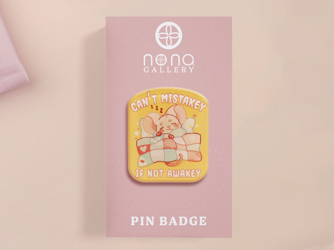 A yellow acrylic pin badge with a pink heart-shaped pin back clasp. The design of the pin badge is of a cute mouse napping in bed with a patchwork quilt with the quote can't mistakey if not awakey.