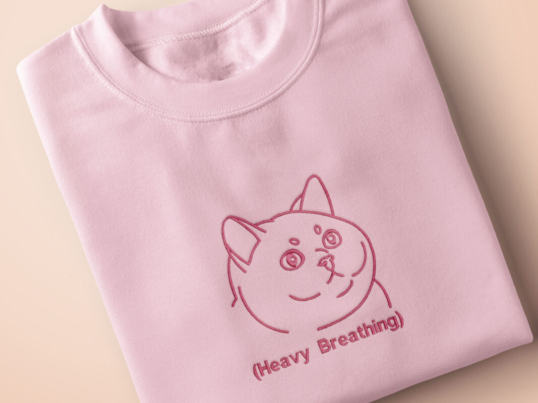 A pink crew neck short sleeve t-shirt, with an embroidered pink thread design of a cute fat cat portrait with text underneath saying (Heavy Breathing)