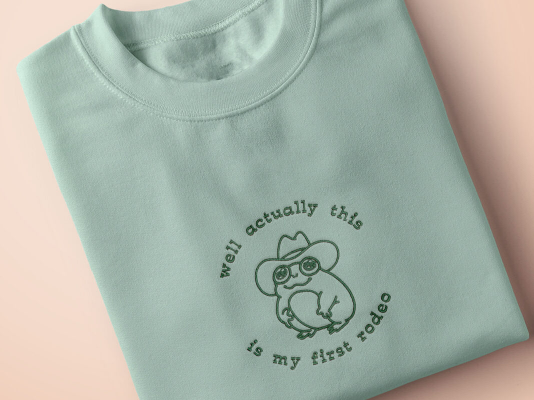 An embroidered green t-shirt design of a cute cowboy frog wearing a stetson surrounded by the test well actually this is my first rodeo.